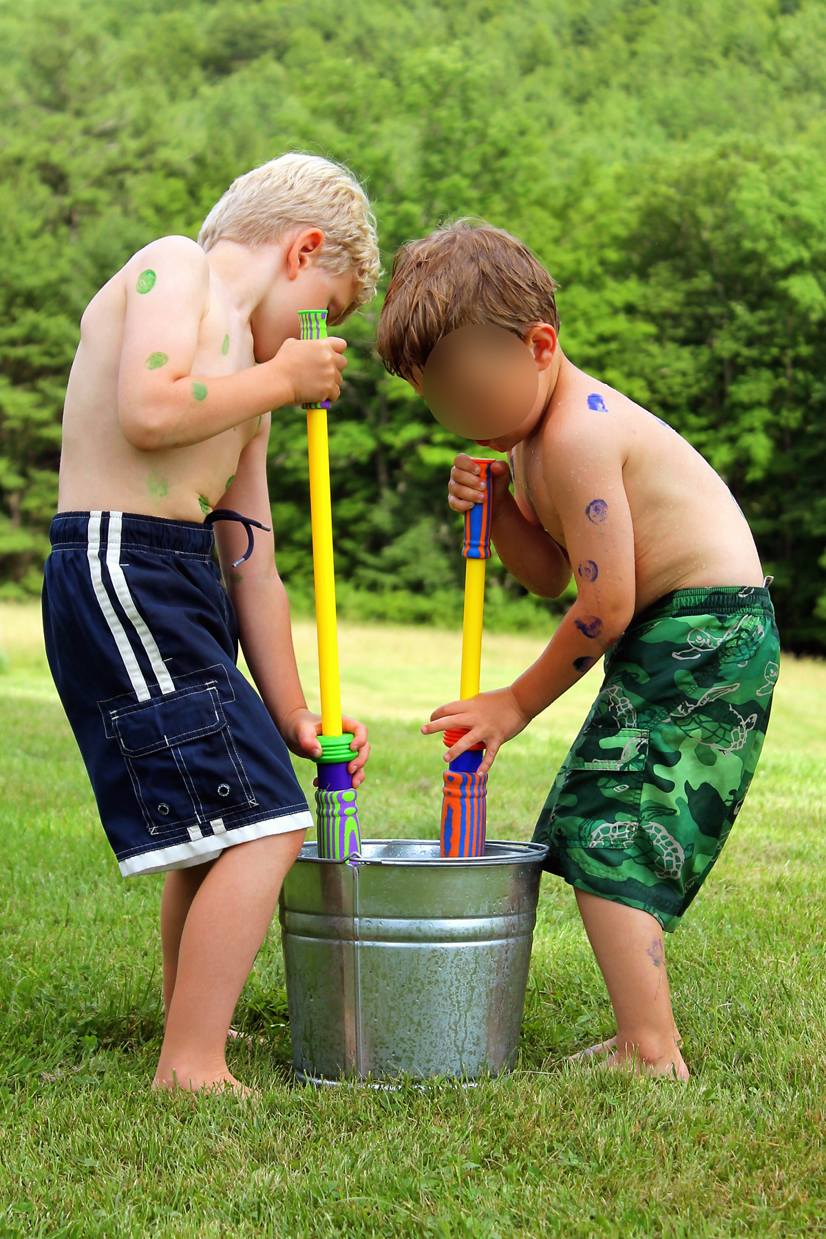 Two boys with paint dots on their skin filling water blasters from a metal bucket during a backyard water game.