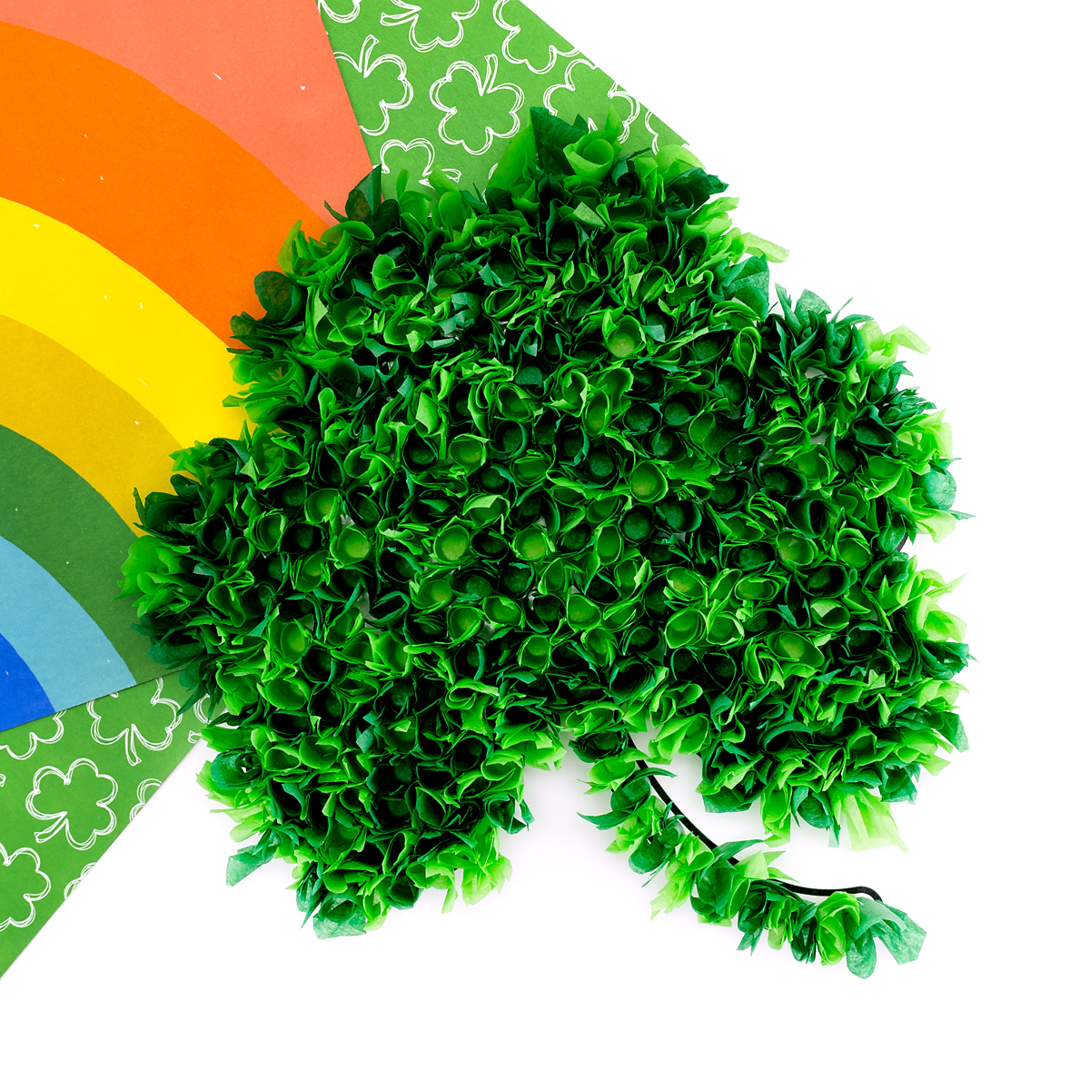 Finished tissue paper shamrock craft on a table with rainbow and shamrock paper. 
