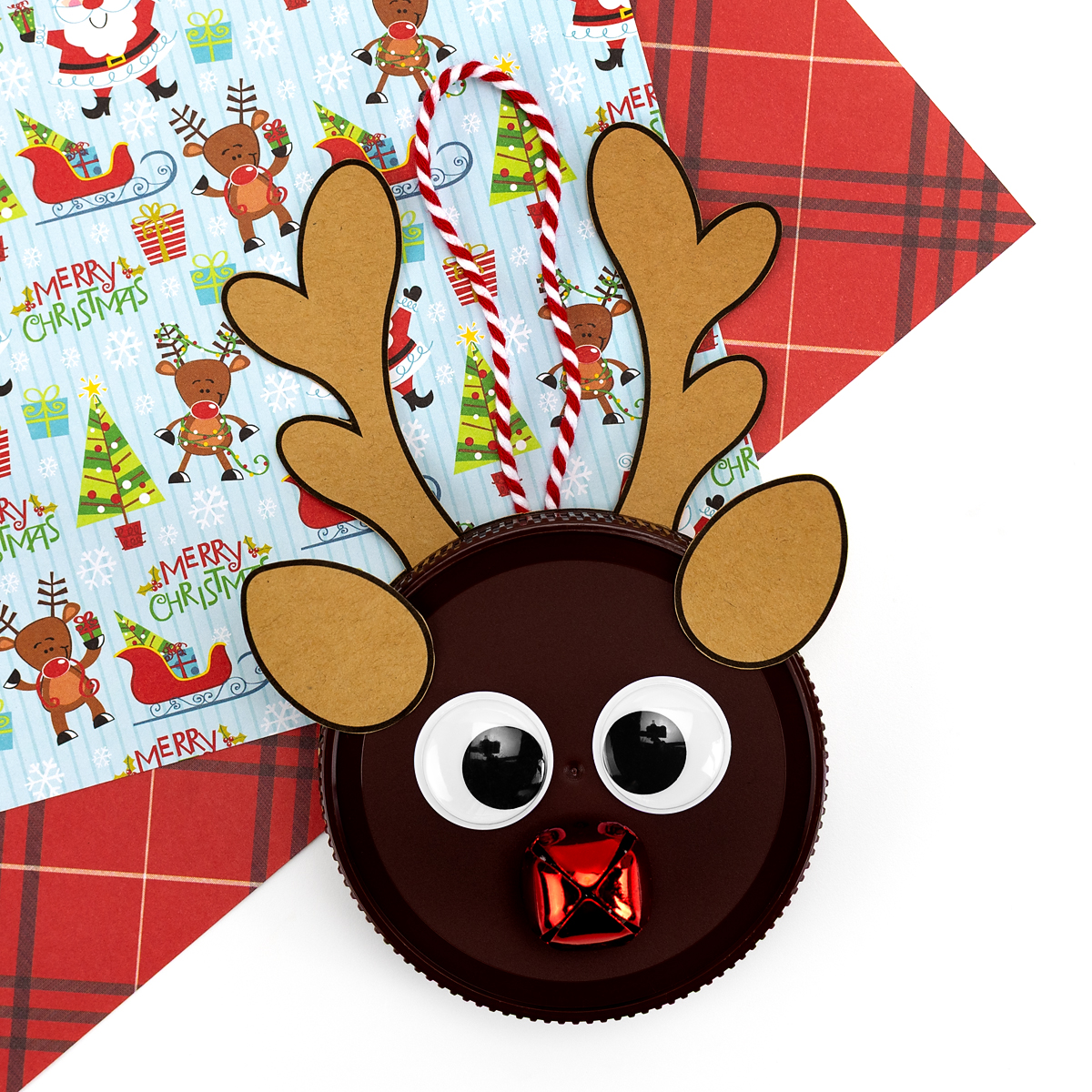 Upcycled jar lid reindeer ornament on a table, decorated with googly eyes, a jingle bell nose, and a string for hanging.