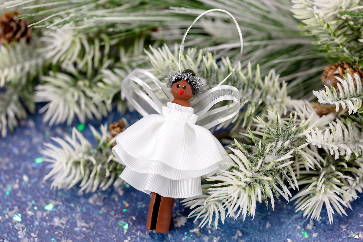 Easy and Fun Christmas Angel Ornament for Kids Finished clothespin angel ornament with black hair, painted face, white ribbon dress, silver wings, and silver halo, perfect for a handmade holiday decoration.