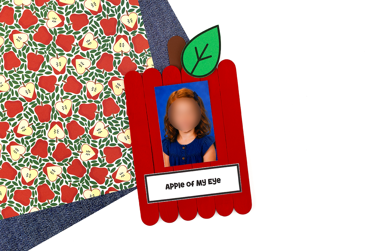 Red popsicle stick apple craft with a felt leaf, brown stem, and a young girl's school photo framed inside, labeled Apple of My Eye.