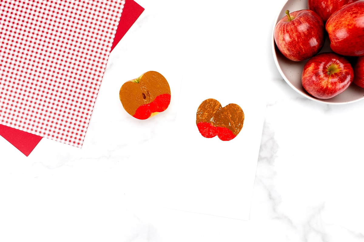 An apple cut in half and painted to make an apple stamp for the caramel apple craft with a bowl of apples in the upper right corner.