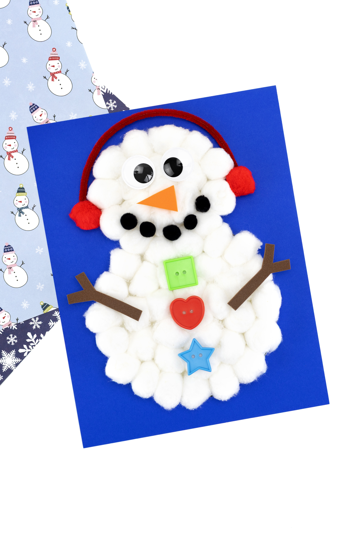 Finished cotton ball snowman on colored cardstock with pom pom earmuffs, carrot nose, pom pom mouth, buttons, and stick arms.