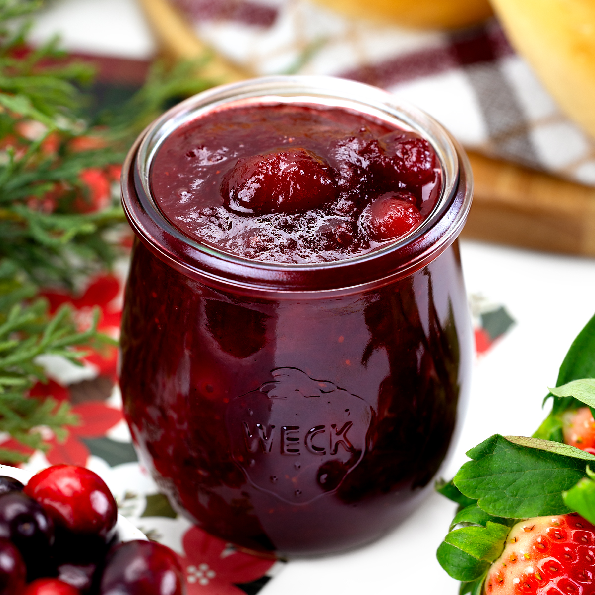 A jar of homemade Christmas Jam with vibrant red strawberries and cranberries, sitting on a wooden board with a spoonful of jam nearby, ready to enjoy or gift.