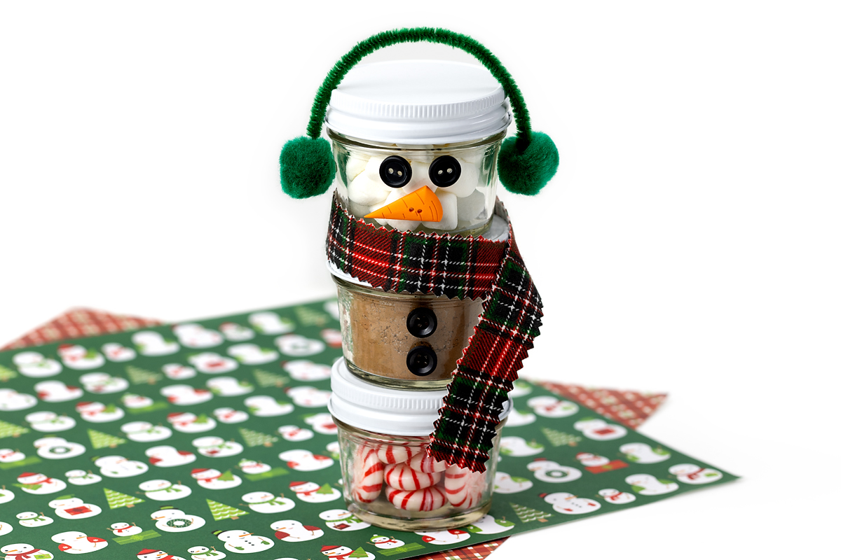 Three stacked mini mason jars forming a snowman, decorated with a flannel scarf and pom-pom earmuffs.