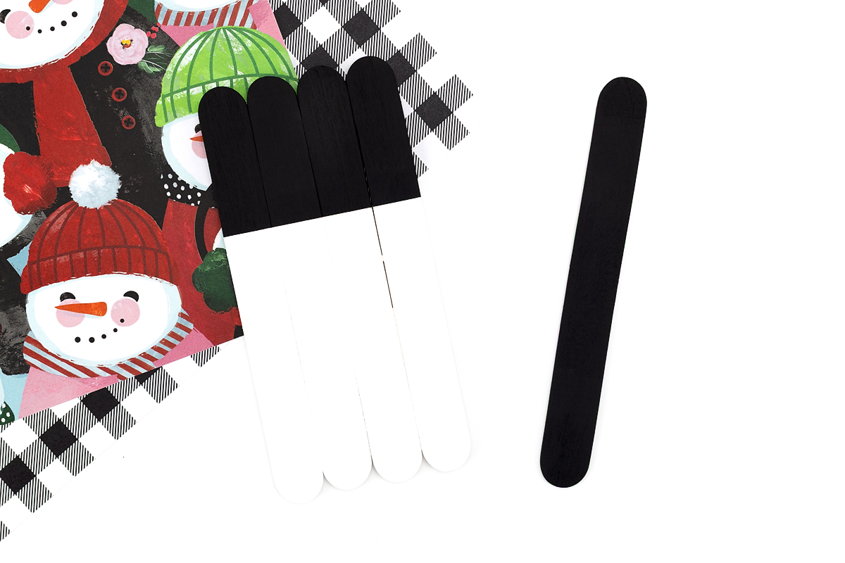 Snowman base painted with white and black acrylic paint next to a black-painted wide popsicle stick for the hat.