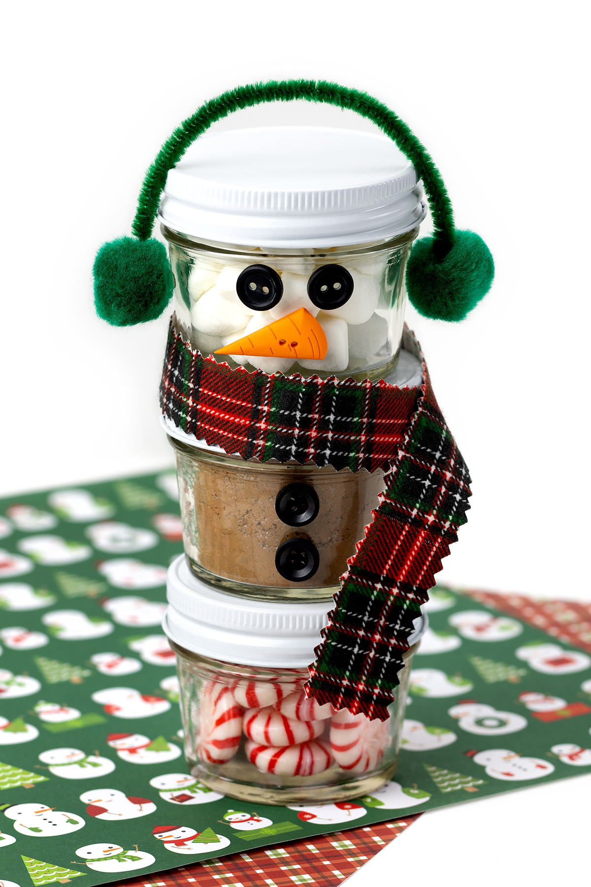 Three decorated mini mason jars stacked to form a snowman, complete with a flannel scarf, pom-pom earmuffs, and button details, ready for gifting.