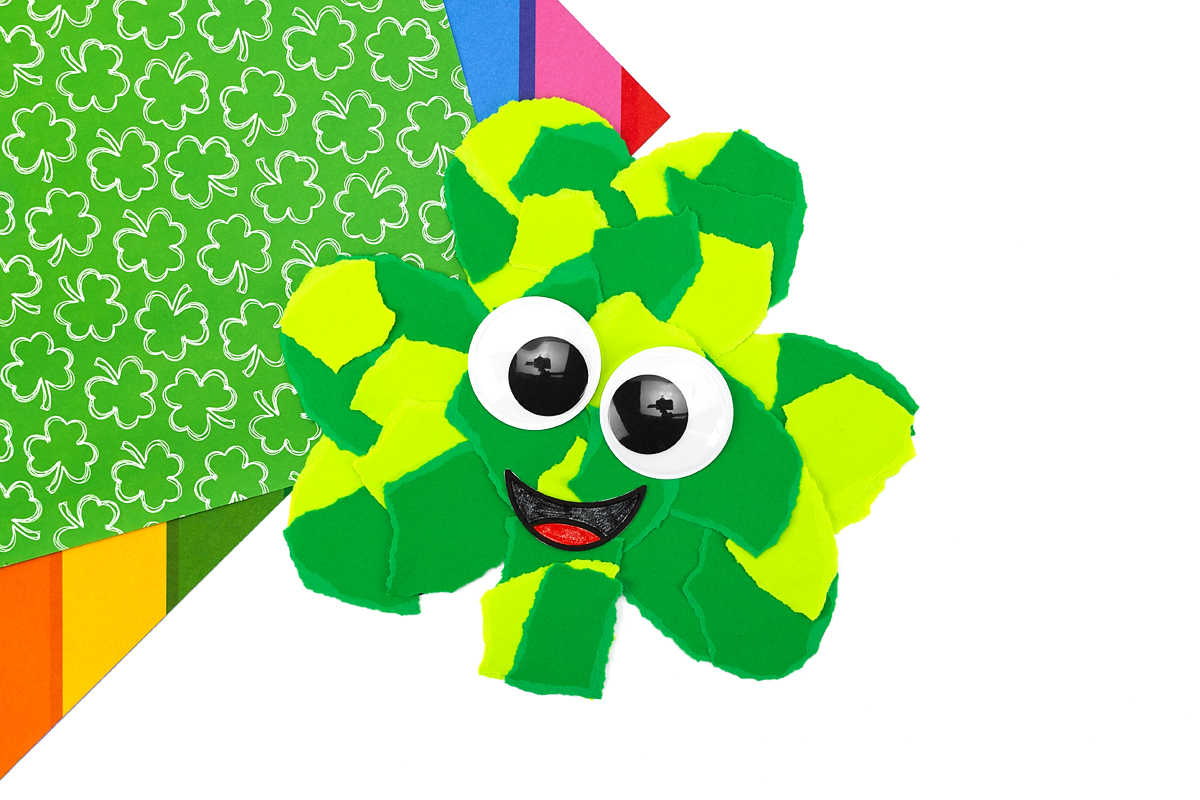 Completed torn paper shamrock craft with googly eyes and smiling face on white background.