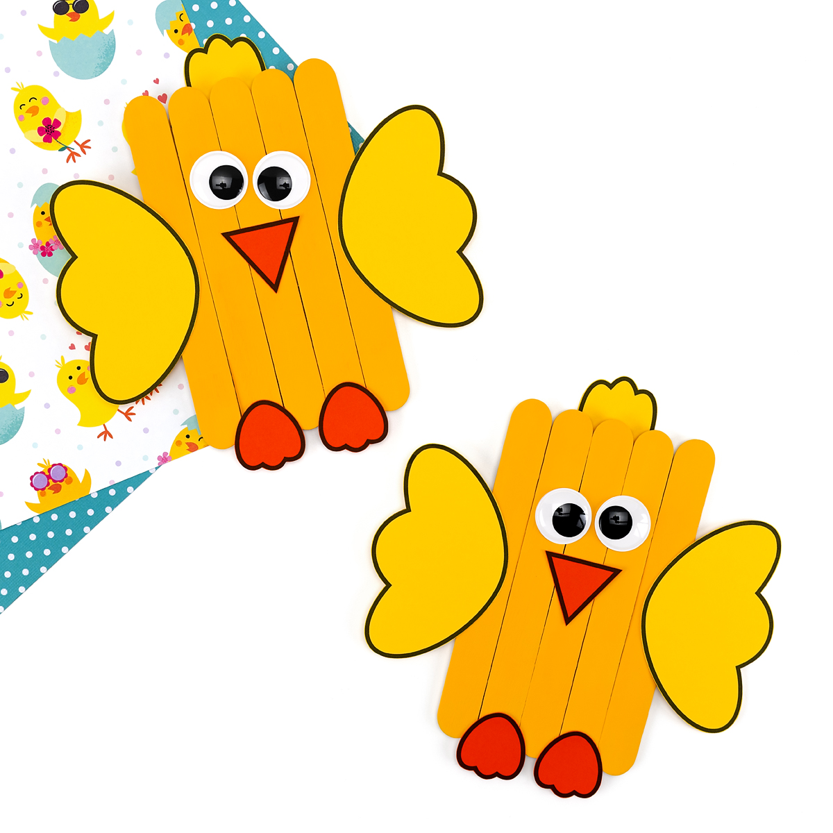 Two finished popsicle stick chicks, decorated with yellow paint, paper wings, tuft of feathers, eyes, beak, and feet, sitting side by side.