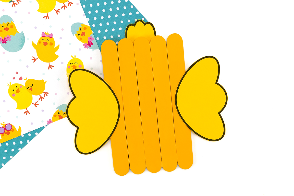 Yellow popsicle stick chick with paper wings glued on the sides and a tuft of paper feathers attached along the top.