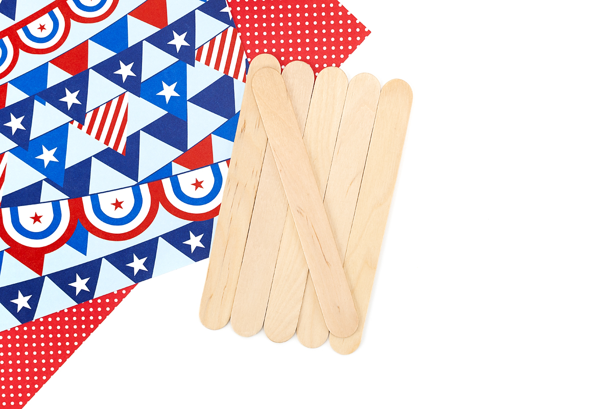 Six craft sticks laid out next to patriotic red white and blue scrapbook paper for an Uncle Sam popsicle stick craft.