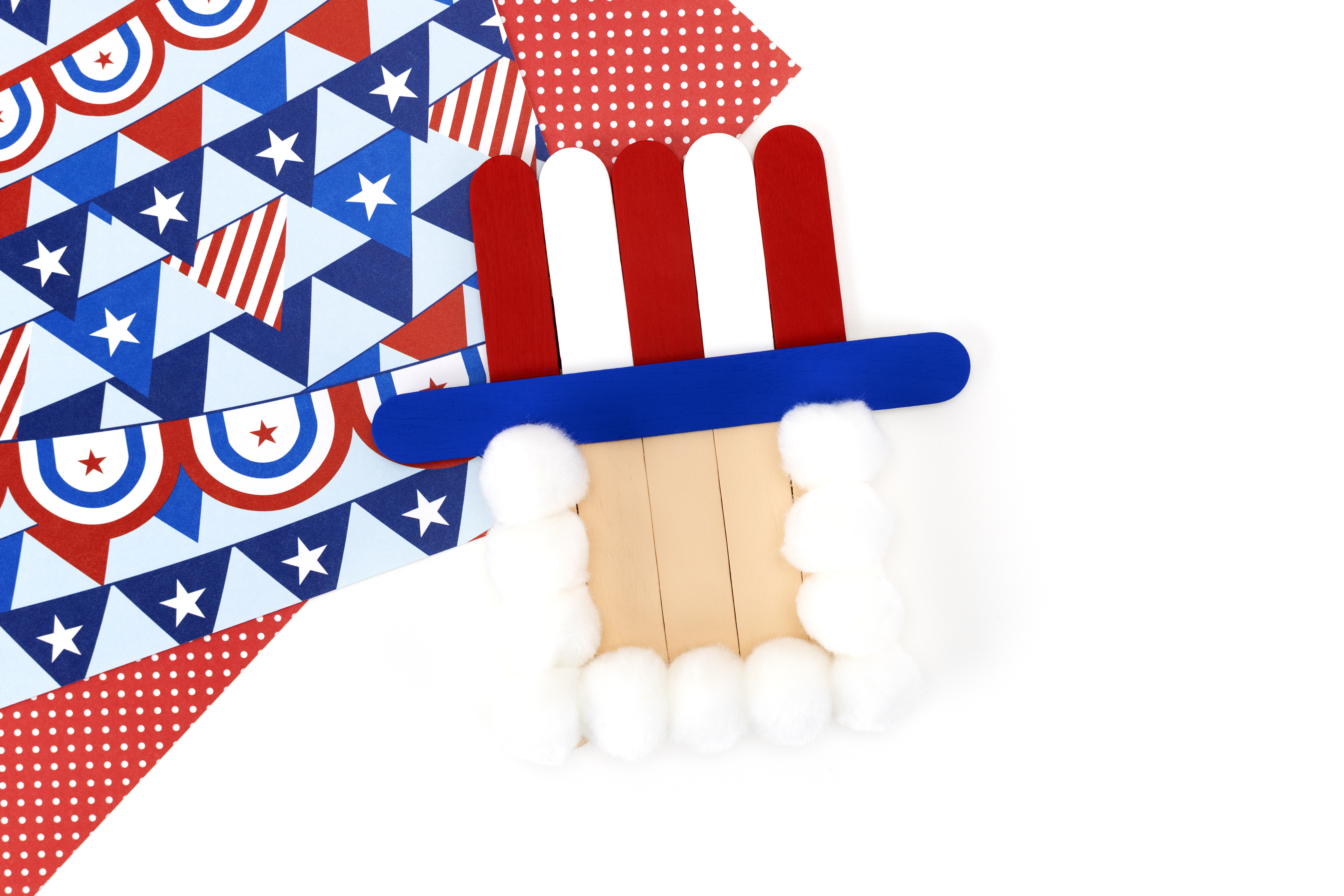 Uncle Sam popsicle stick craft in progress with white pom poms glued around the face to form a beard.