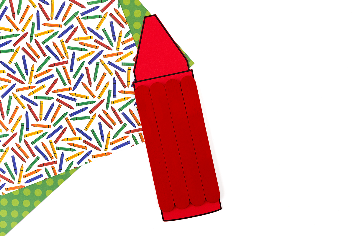 Finished red popsicle stick crayon craft with pointed tip and rounded bottom attached, displayed on a colorful crayon-print background.