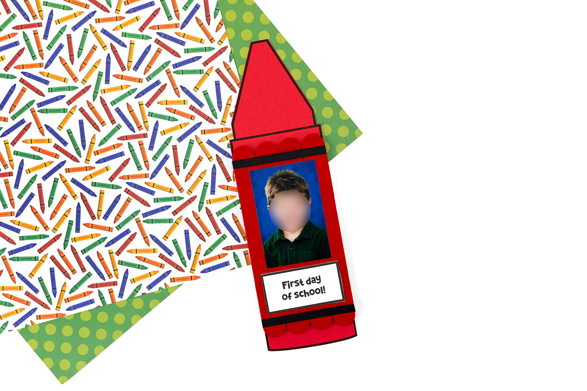 Red popsicle stick crayon craft with a school photo in the center and a "First Day of School" label at the bottom, displayed on a crayon-print background.