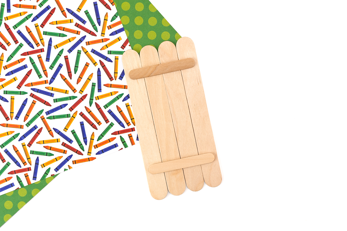 Four unpainted jumbo popsicle sticks aligned side by side with two regular popsicle sticks hot glued horizontally across the back to form the base of a crayon craft.
