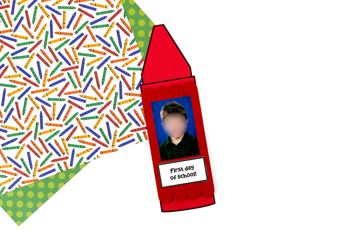 Single finished red popsicle stick crayon craft with a school photo and "First Day of School" label, displayed on a colorful crayon-print background.