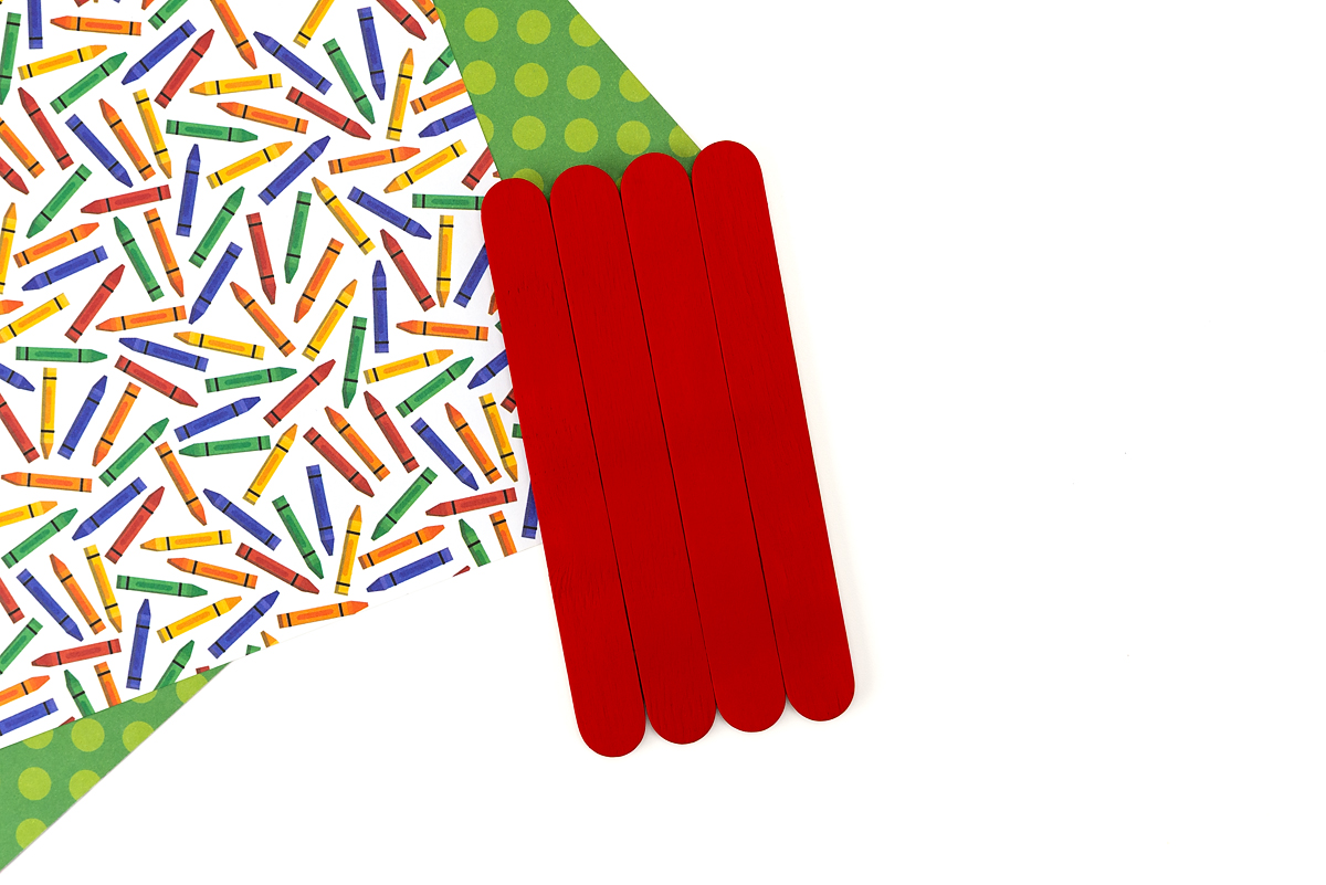 Four red-painted jumbo popsicle sticks laid side by side on a colorful crayon-print background, ready to be assembled into a crayon craft.