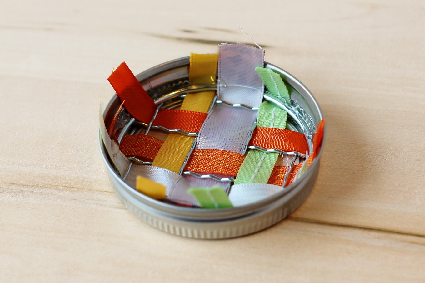 Just in time for fall, learn how to make vibrant pumpkin suncatchers with scraps of woven ribbon and mason jar lids. This weaving craft is perfect for kids!